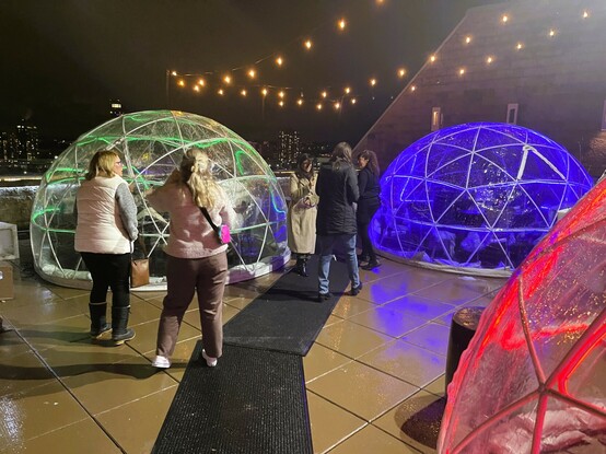A picture featuring the large clear "igloos" at Over the Charles rooftop bar deck. One has green neon lighting, one blue and one red. They are about 8' high in the middle and probably have a width of about 15 feet. 