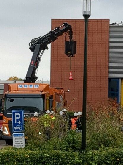 A truck with Hydraulic Crane lifting a traffic cone. Seven workers with safety gear look on.