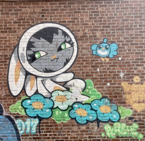 Wall mural in old Montreal on red brick wall. A creature wearing a white one piece bunny costume, sitting in a patch of flowers.
